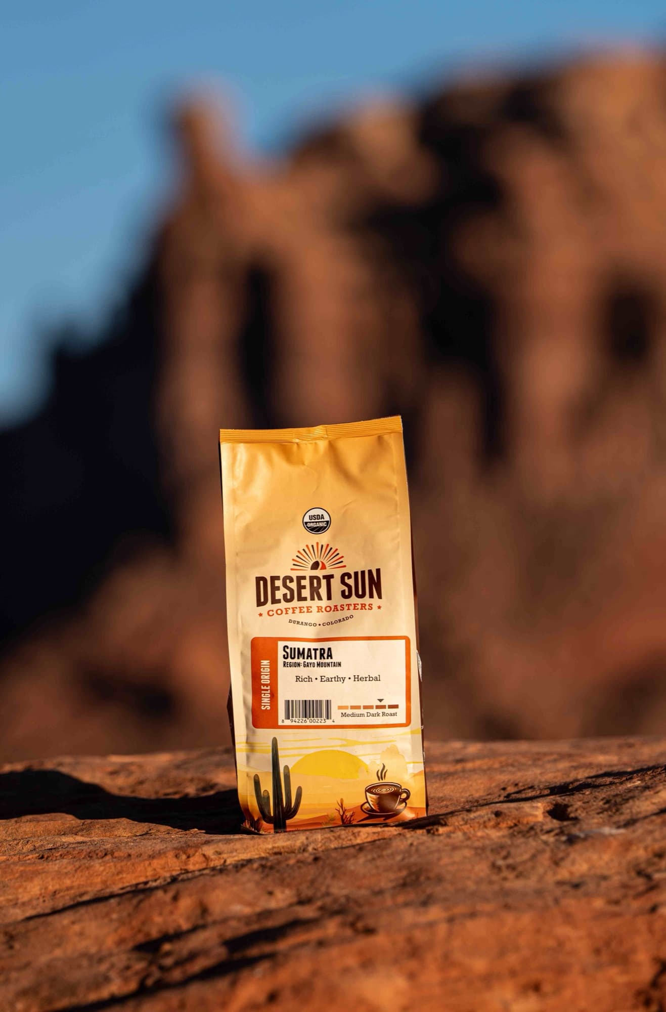 A Desert Sun coffee bag standing on red rock with a sandstone tower behind