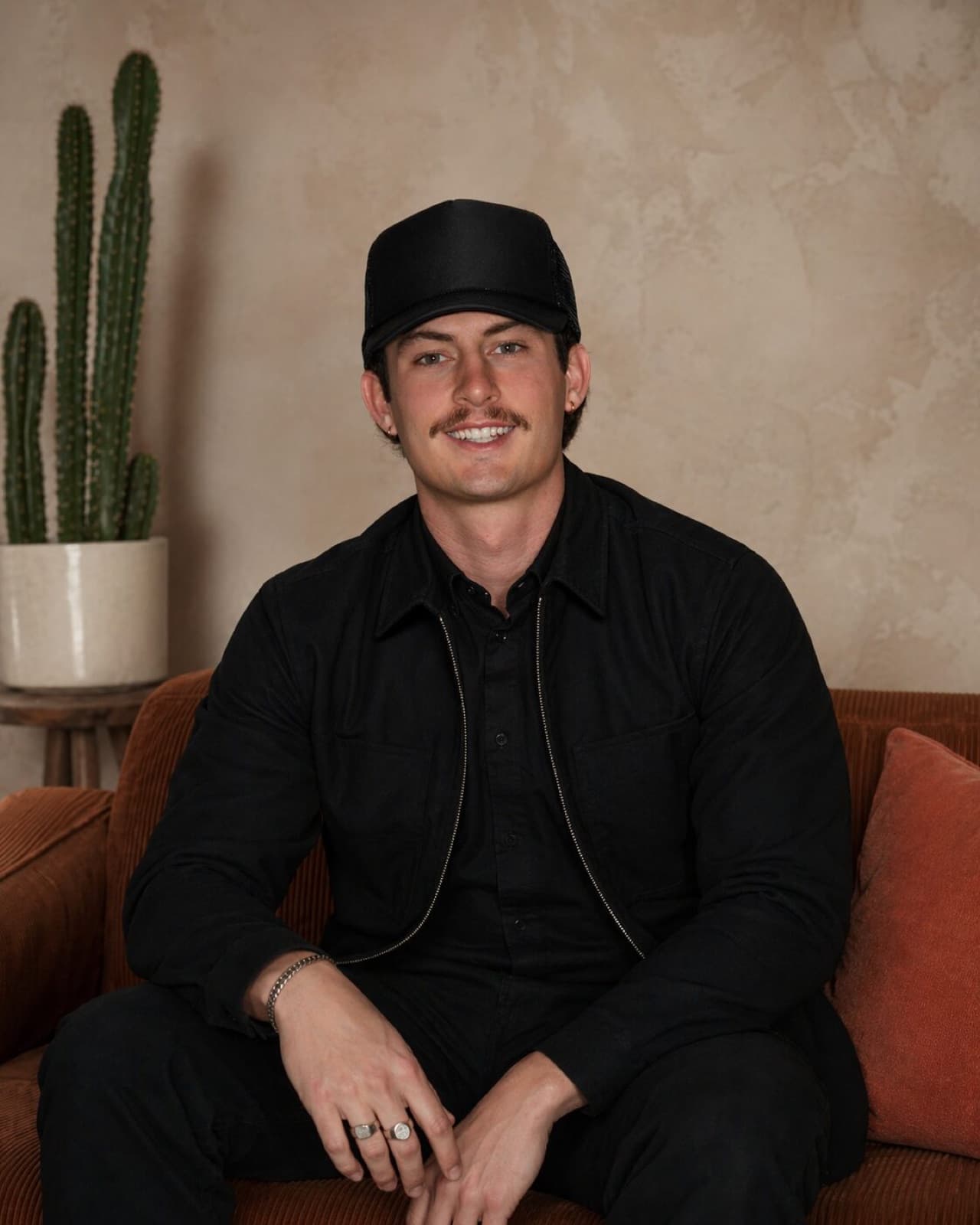 Kahlil Ballenger, in a black cap, on a rust corduroy sofa beside a tall cactus