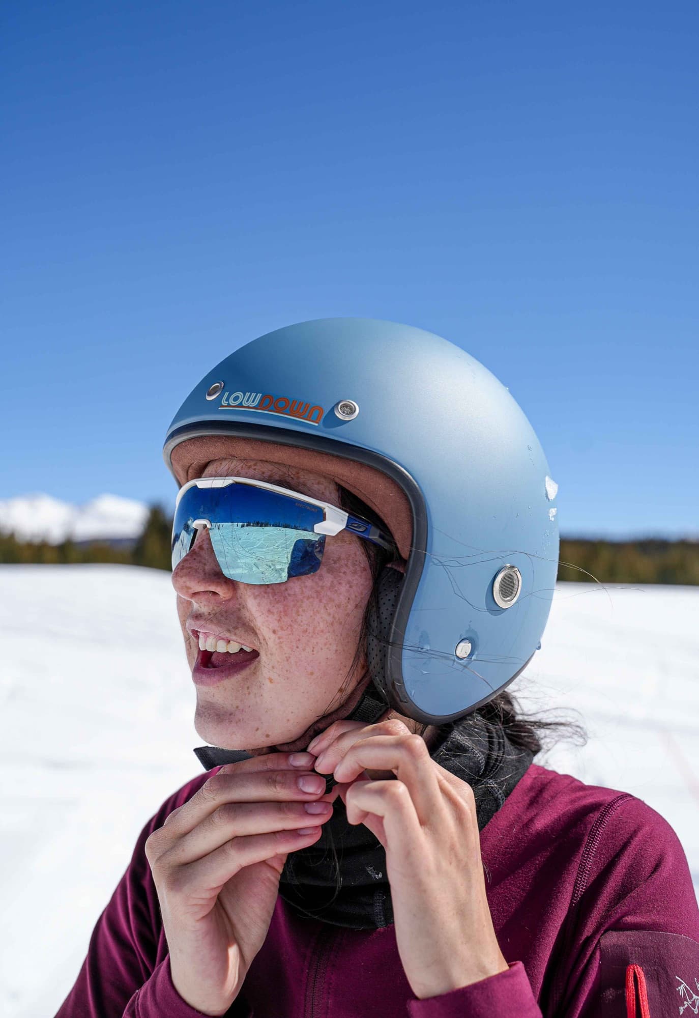 A skier fastens the strap of a pale blue Lowdown helmet, peaks behind