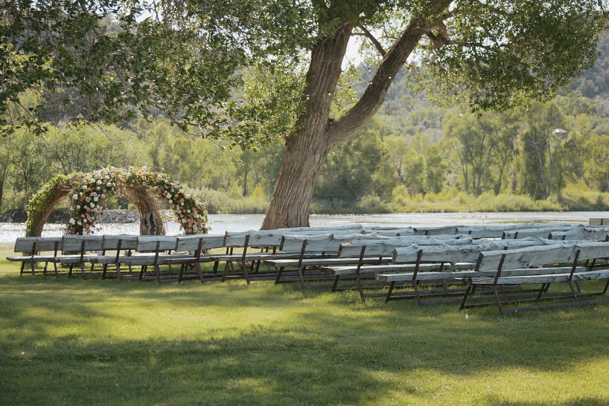Wooden ceremony benches under a cottonwood facing a floral arch beside the pond
