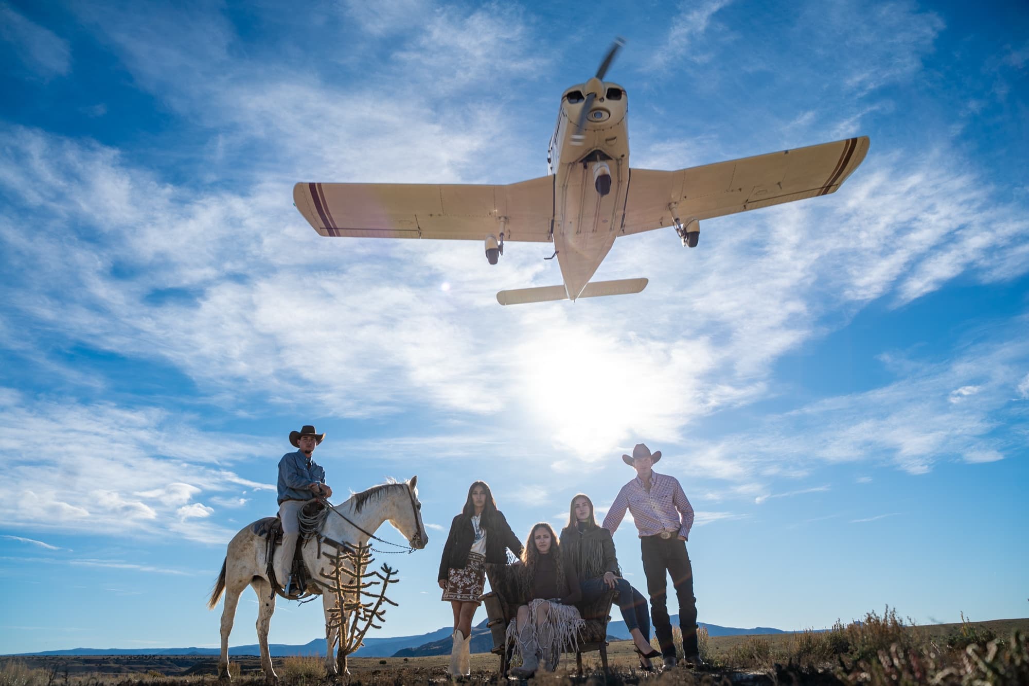A small plane passes low over four riders in western wear on a dirt road